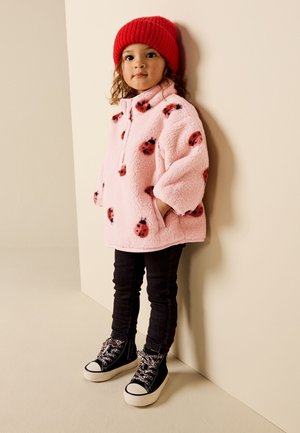 Bambino con cappello rosso di lana, giacca in pile rosa con stampe di coccinelle, pantaloni neri e sneaker alte nere, in piedi contro un muro beige.