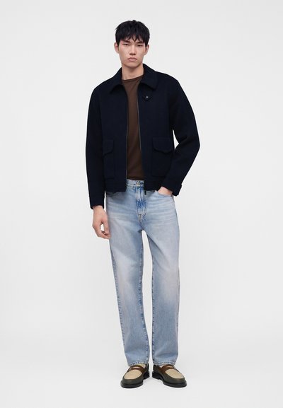 Jeune homme portant une veste noire, une chemise marron, un jean bleu clair et des chaussures beiges et noires, debout sur un fond blanc.