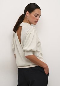 Pulover tricotat de culoare bej deschis, cu decolteu în formă de V la spate, mâneci puff și talie ajustată. Material sclipitor, cu textură. Purtați-l cu pantaloni închis la culoare.