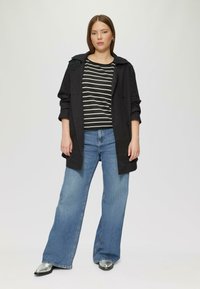 Manteau noir sur une chemise à rayures noir et blanc, associé à un jean large bleu et des chaussures argentées à bouts pointus. Design simple et texturé.