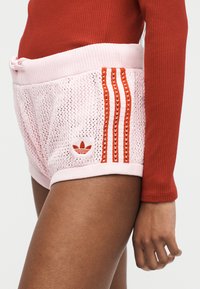 Άτομο που φοράει ροζ πλεκτό σορτς με κόκκινες ρίγες και το λογότυπο της Adidas, συνδυασμένο με μακριμάνικη κόκκινη ριμπ μπλούζα, εμφανιζόμενο από μέση έως μέση-μηρού.