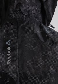 Czarna kurtka Reebok z teksturową powierzchnią, z delikatnym geometrycznym wzorem i logotypem. Regulowany kaptur i lekki materiał.