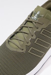 Olivgrön träningsskor med nätöverdel, strukturerad design, reflekterande logotyp och platta snören. Vit sula med en mörk gummitå.