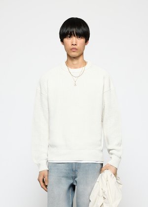 Young man with black hair wearing white textured sweater and light blue jeans, holding a white jacket in his left hand.