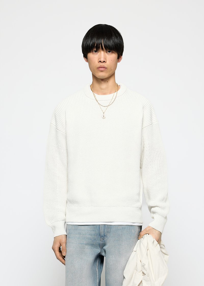 Junger Mann mit schwarzen Haaren, der einen weißen strukturierten Pullover und hellblaue Jeans trägt, während er eine weiße Jacke in seiner linken Hand hält.