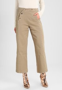 Mujer con pantalones de talle alto color beige con detalles de botones a presión, una mano en el bolsillo, combinados con botas de tacón ancho metálicas color rosa dorado.