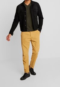 Schwarze Jeansjacke über einem dunkelgrünen V-Ausschnitt-Shirt, kombiniert mit gelben Cordhosen und schwarzen Sneakern. Lässiger Schnitt mit strukturierten Materialien.