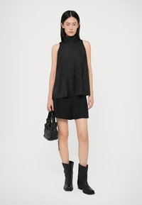 Vrouw in een mouwloze zwarte top met hoge hals, zwarte shorts en zwarte laarzen tot halverwege het kuit, die een kleine zwarte tas met studs vasthoudt.