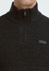 Mørkegrå, struktureret sweater med høj krave og halv lynlås. Har et mærke foran nær kraven.