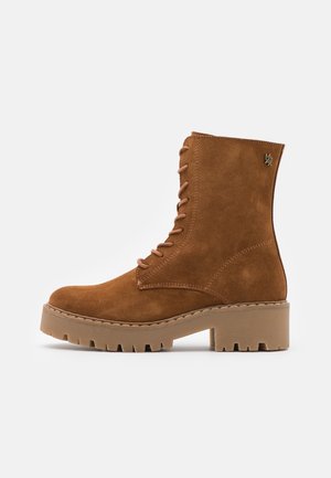 Bottine marron en daim à lacets avec semelle crantée épaisse et petit talon carré, ornée d'un petit logo métallique de la marque sur le côté supérieur.