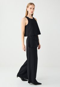 Schwarzes ärmelloses Oberteil mit schichtweise Gestaltung, kombiniert mit weit geschnittenen schwarzen Hosen. Glattes Material mit minimalen Akzenten, taillierte Passform.