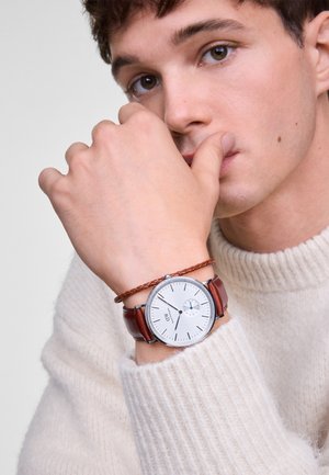 Montre ronde en argent avec un cadran blanc et deux accents bleus, montée sur un bracelet en cuir marron avec un détail tressé. Portée sur la manche d'un pull en tricot.