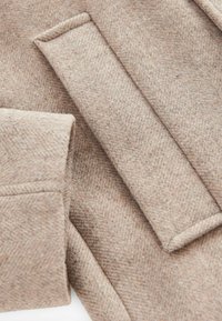 Béžový vlněný kabát s herringbone texturou, s sklápěcími manžetami a klopami. Materiál vyzerá měkko a teplo, zdůrazňuje komplikované tkaní.