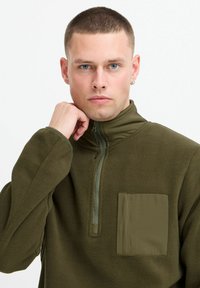 Olive green fleece pullover with a high collar, quarter-zip front, and a square pocket on the left chest. Soft texture, casual design.