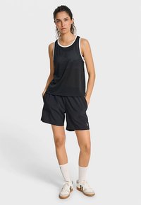 Sort mesh tanktop med hvid kant, kombineret med løse sorte shorts. Modellen har hvide sneakers og hvide knæstrømper på.