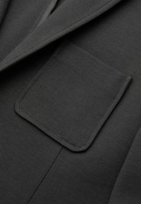 Primer plano de una chaqueta de tela gris oscuro que muestra un bolsillo rectangular con costura detallada cerca de la solapa.