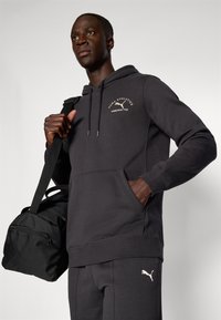Sort hættetrøje lavet af blødt stof, med en kængurulomme og snor i hætten. Puma-logo i lysegrå på brystet og bukserne. Sort sportsduffel bag.