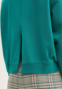 Sukienka w kolorze teal z rozcięciem z tyłu i zaokrąglonym dołem; zestawiona z spodniami w kratę w odcieniach beżu, czerni i zieleni.