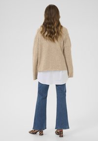 Beige Strickpullover mit lockerer Passform, über einem weißen Hemd getragen. Kombiniert mit blauen, ausgestellten Jeans und Hausschuhen mit Animal-Print und Absatz.