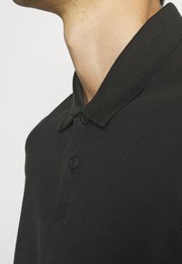Svart polo skjorta med en texturerad krage, med två knappar och en slim fit-design. Tyget verkar slätt och något elastiskt.