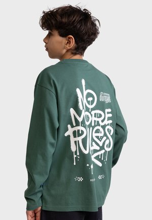 Dreng med mørkt krøllet hår iført en grøn langærmet skjorte med "No More Rules" i hvid graffiti tekst på bagsiden.