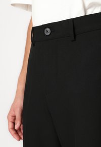 Κοντινής λήψης μαύρων παντελονιών με κουμπωμένη μέση, φορεμένα με λευκό πουκάμισο, που δείχνουν την πλαϊνή τσέπη και μέρος του χεριού του ατόμου.