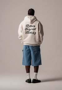 Krém kapucnis pulóver hátul fekete, dőlt betűs "Blkvts Original Clothing" felirattal, kék farmer rövidnadrággal és fekete cipővel párosítva.