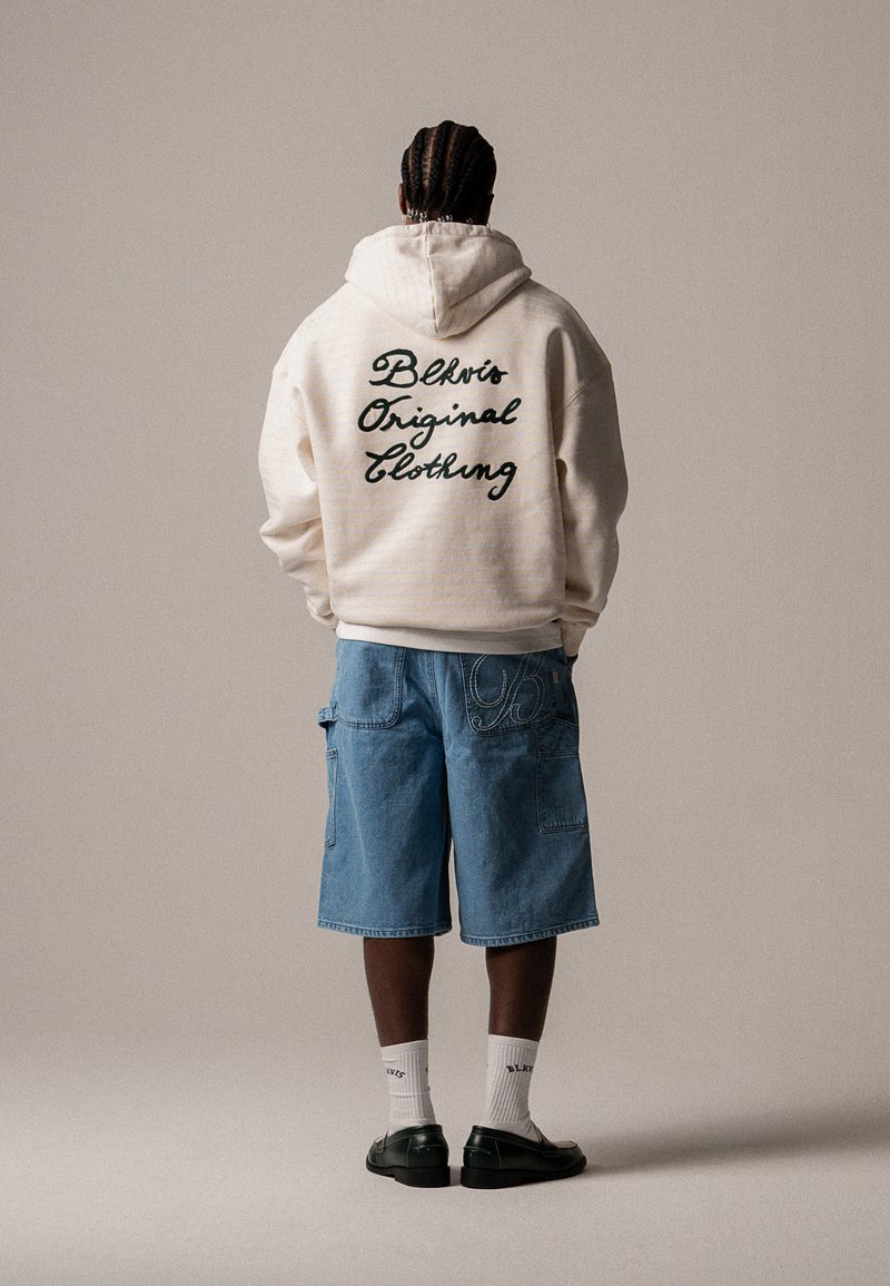 Krém kapucnis pulóver hátul fekete, dőlt betűs "Blkvts Original Clothing" felirattal, kék farmer rövidnadrággal és fekete cipővel párosítva.