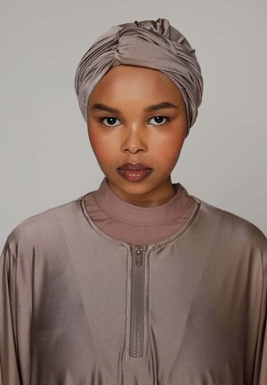 Femme à la peau mate portant un turban en satin taupe et un haut zippé assorti, face à l'avant avec une expression neutre sur un fond uni.