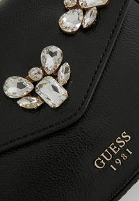 Portefeuille Guess 1981 en cuir texturé noir avec des ornements en cristal doré de différentes formes.