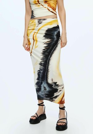Midi sukňa s abstraktným čiernym, žltým a hnedým tie-dye vzorom, nosená s zodpovedajúcim crop topom a čiernymi platformovými sandálmi s remienkami okolo členkov.