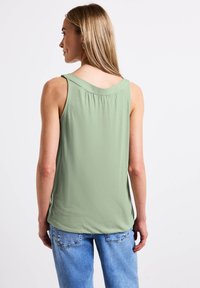 Bezramenný top v světlezelené barvě, s kulatým výstřihem a volným střihem, kombinovaný s modrými džíny. Měkká textura a jednoduchý design.