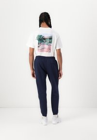 T-shirt branca com um gráfico colorido nas costas, combinada com calças de treino azuis marinho. Ténis completam o visual casual.