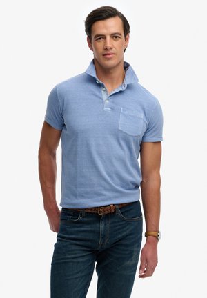 Mann trägt hellblaues kurzärmeliges Poloshirt mit Brusttasche, dunkle Jeans, braunen geflochtenen Gürtel und Uhr, steht vor weißem Hintergrund.