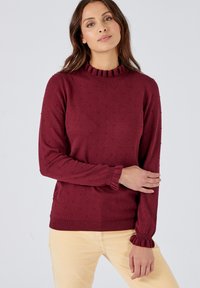 Maglione a maglia bordeaux con un motivo testurizzato, colletto alto a coste e maniche svasate. Il materiale sembra morbido e accogliente.