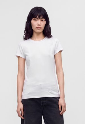 Kvinde med langt sort hår iført en ensfarvet hvid kortærmet t-shirt og mørke bukser, stående mod en lysegrå baggrund.