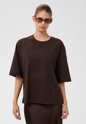 Jeune femme portant un t-shirt marron foncé oversize, une jupe assortie et des lunettes de soleil teintées ambre, debout devant un fond clair uni.