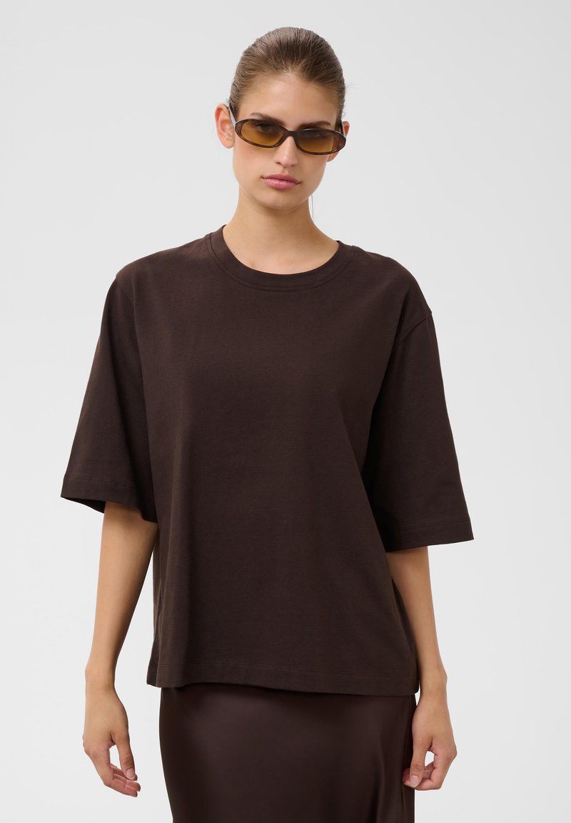 Jeune femme portant un t-shirt marron foncé oversize, une jupe assortie et des lunettes de soleil teintées ambre, debout devant un fond clair uni.