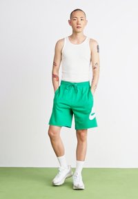Shorts en coton verts avec une taille élastique et un cordon de serrage, ornés d'un logo Nike blanc, associés à un débardeur blanc et des baskets blanches.