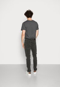 Hombre con cabello rizado vestido con una camiseta gris oscuro, jeans negros y zapatillas blancas, de pie frente a una pared blanca con las manos relajadas.