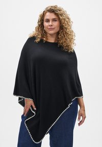 Črn pončo s širokim, razkošnim dizajnom in valovitim belim robom na dnu. Izdelan iz mehke tkanine, nošen čez modre kavbojke.