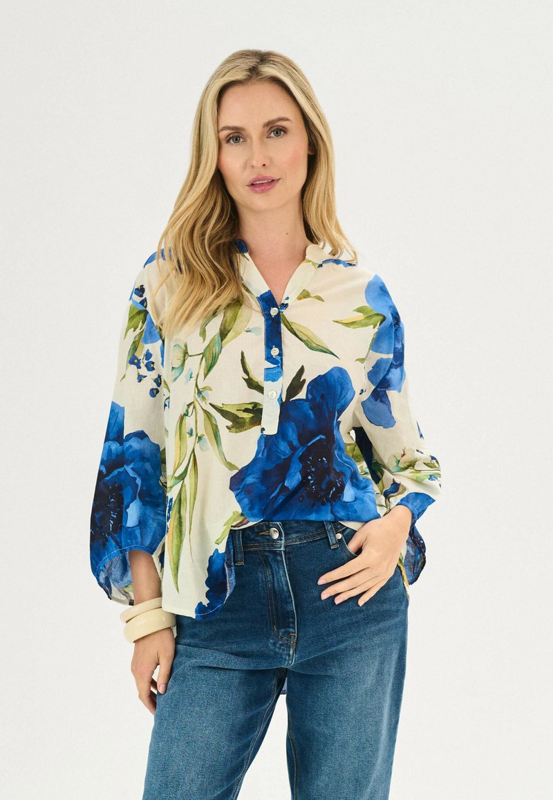 Une chemise à boutons florale présente des fleurs bleues et vertes sur un tissu beige, avec un col et des manches longues légèrement évasées.