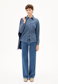Denim overhemd met knoopsluiting, zakken en lange mouwen. Gecombineerd met wijde blauwe jeans en beige schoenen. Achtergrond is simpel wit.