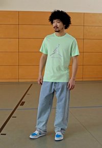 T-shirt in cotone verde chiaro con logo Jumpman, abbinata a jeans baggy blu chiaro e sneakers blu chiaro. Sfondo semplice.