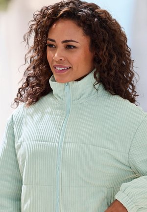 Woman with curly hair wearing a light blue ribbed zip-up jacket, smiling and looking slightly right against a bright background.