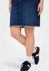 Temno moderna denim krila s raztrganim robom, v kombinaciji z mornarskimi supergami z belimi akcenti in belimi nogavicami. Položaj nog je prekrižen.