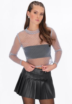 Jonge vrouw met lang haar, gekleed in een doorschijnende skinny top met polkadots, een donker bandeau en een zwarte geplooide leren rok, die haar tailleband rechtzet.