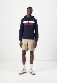 Felpa della marina con strisce bianche e bordeaux, tasca a marsupio e coulisse. Indossata con pantaloni corti beige e calze bianche, completata da sneakers chiare.