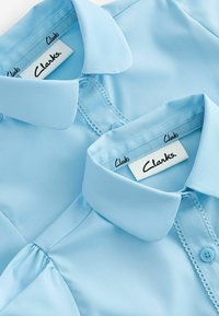 Camicia col cielo chiaro con colletto, caratterizzata da chiusura con bottoni e cuciture decorative. Due camicie sovrapposte con etichette Clarks visibili all'interno.