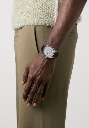 Montre en argent avec un cadran gris, un bracelet en caoutchouc noir et des accents argentés. Portée sur une main sur un tissu de pantalon vert olive.